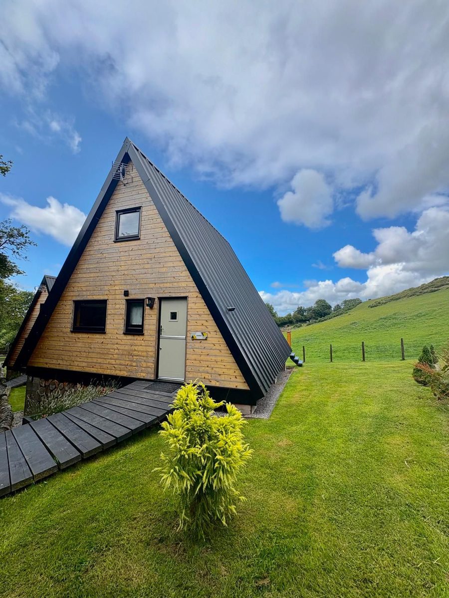 A-frame timber holiday lodge with a decked path, set against a green Welsh hillside